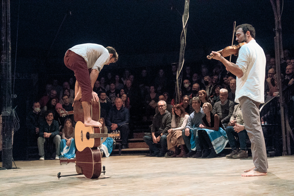 Ein Akrobat des Le P'tit Cirk führt in der Manege ein akrobatisches Balance-Kunststück auf Gitarren auf. Ein anderere Akrobat begleitet ihn dabei auf der Geige.
